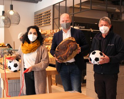 Peter Ostendorf von mel&koffie, Sya Ahmad, Bezirksvorsteherin neunter Bezirk und Georg Jelenko, Leiter Samariterbund Sozialmärkte in mel&koffie Filiale im neunten Bezirk, halten Fußbälle, Springseile und einen Laib Brot in der Hand