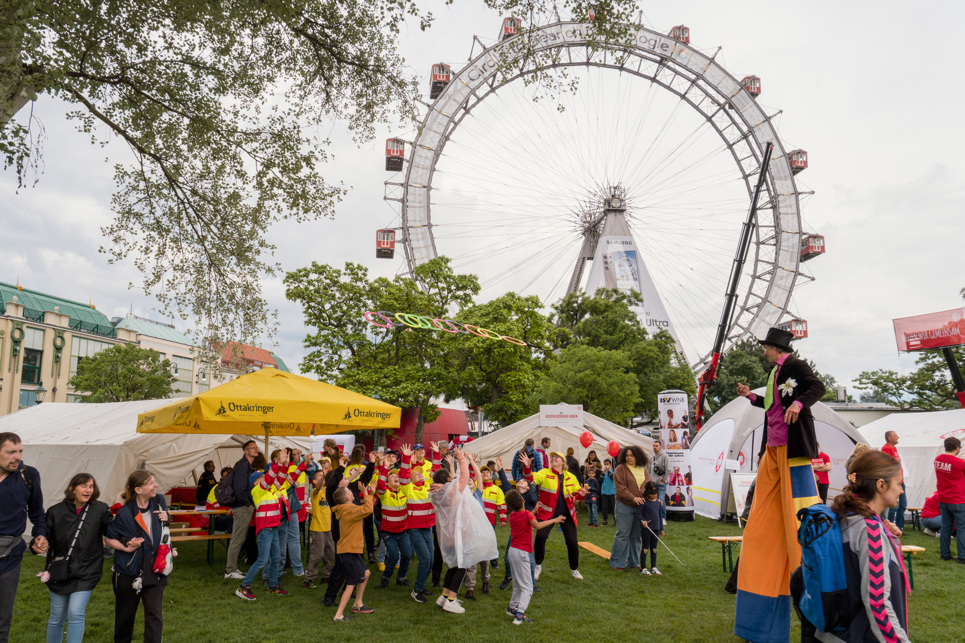 Aufnahme vom Tag des Samariterbundes, im Vordergrund Stelzengeher, der Ringe zu Kindern wirft, im Hintergrund Riesenrad, mehrere Schirme, Zelte und Menschen