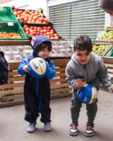 zwei kleine Buben in Sozialmarkt (im Hintergrund Kisten mit Obst), spielen mit Bällen, Bub rechts versucht Ball wie Basketball zu drippeln, Bub links hält Ball in beiden Händen und schaut zu