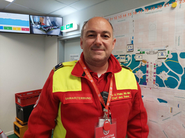 Ein Mann in einer roten und gelben Uniform mit dem Aufdruck „Samariterbund“ steht in einem Raum. Im Hintergrund sind eine Karte und ein Monitor sichtbar. Der Mann lächelt und trägt einen Ausweis um den Hals.