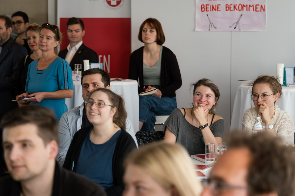 Auf dem Bild sind mehrere lächelnde Personen zu sehen, die um runde Tische sitzen.