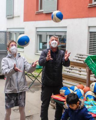 Georg Jelenko und Bezirksvorsteherin 15. Bezirk in Sozialmarkt Pillergasse, werfen Bälle in die Luft, tragen beide eine FFP2-Maske, im Vordergrund Kind, dass auf weitere Artikel blickt, im Hintergrund Kisten mit Lebensmitteln