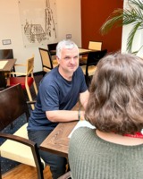 Mann mit dunkelblauem T-Shirt und grauen Haaren sitzt an Tisch und blickt direkt in Kamera, daneben Person von hinten fotografiert, sieht nur Schulterbereich und Hinterkopf, sitzen in einem Raum, dahinter weitere Tische und Stühle, Pflanzen, Einrichtung