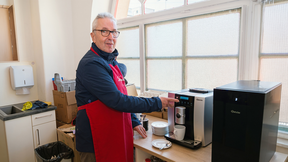 Ein Mann mit Brille und Schürze steht in einer hellen Küche und bedient eine Kaffeemaschine. Auf dem Tisch sind eine Tasse und einige Gegenstände sichtbar, während der Mann lächelt und auf die Maschine zeigt.