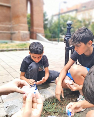 Vier Kinder, die im Freien auf dem Boden sitzen und ein Experiment mit einem Messbecher und einer Pipette durchführen.