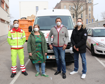 vier Personen, alle tragen FFP2-Maske, einer davon (ganz links) in Samariterbund Uniform, stehen vor einem Kleintransporter, im Hintergrund oranger Container, Gebäude