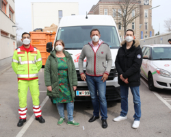 vier Personen, alle tragen FFP2-Maske, einer davon (ganz links) in Samariterbund Uniform, stehen vor einem Kleintransporter, im Hintergrund oranger Container, Gebäude