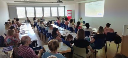 Aufnahme während eines Seminars, vorne eine Vortragende vor Leinwand, viele Teilnehmer:innen auf Stühlen, die zuhören