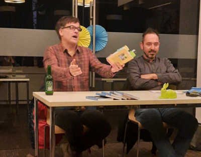 zwei Männer, links Bernd Watzka mit gelbem Buch in der Hand, rechts Bernd Remsing, sitzen an einem Tisch vor einer Glaswand, Internetcafé, ZwischenSchritt, Lesung,