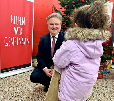 Michael Ludwig kniet vor Mädchen mit Winterjacke, dass man nur von hinten sieht, sie hält ein Geschenk in der Hand, im Hintergrund Roll up mit Slogan Helfen wir gemeinsam und Christbaum