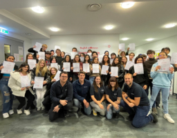 Gruppenfoto mit vielen Personen, halten Zertifikate in der Hand, davor kniend Trainer:innen in blauen Polos mit Samariterbund Logo davor 