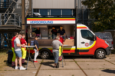 Aufnahme Foodtruck im Samariterbund Autobranding, am oberen Rand des Wagens Schriftzug mit Versorgung, davor mehrere Personen, die sich anstellen oder an Tischen mit Essen stehen