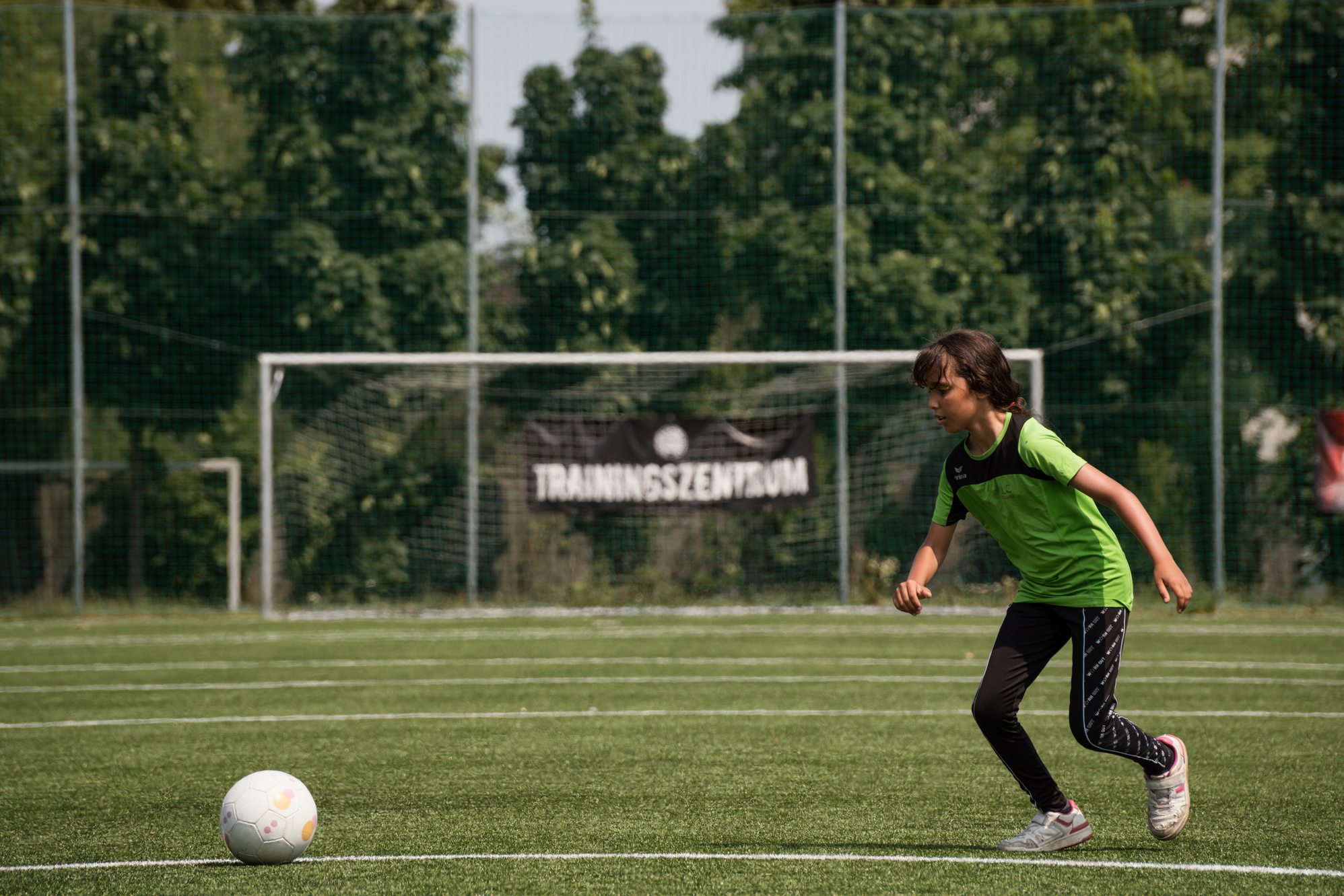 Mädchen auf Fußballfeld, läuft in Richtung eines Fußballs, im HIntergrund Tor erkennbar