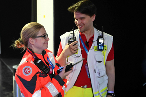 Zwei Mitarbeiter im Rettungsdienst stehen in einem Innenraum. Die Frau trägt eine rote Rettungsjacke und hält ein Funkgerät. Der Mann trägt eine weiße Weste und lacht, während er ebenfalls ein Funkgerät in der Hand hält. Beide sehen entspannt aus und sind in einem Gespräch vertieft.