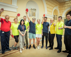 Gruppenfoto bei Tischtennisturnier im Hotel de France, Ukrainische Flüchtlinge und Betruer:innen, umarmen sich am Rücken und halten Tischtennisschläger in die Luft, alle blicken in Kamera und jubeln