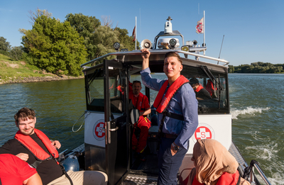 Erik von Satansbratan auf Boot der Wasserrettung vom Samariterbund Wien, blickt in Kamera und lächelt, daneben sitzen weitere Menschen mit Sicherheitswesten auf Boot