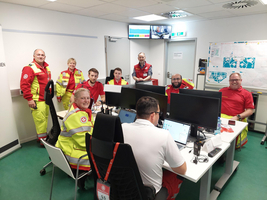 Eine Gruppe von Rettungsdienstmitarbeitern sitzt und steht in einem hellen Raum. Einige tragen rote und gelbe Einsatzkleidung. Am Tisch stehen Computer, während die Mitarbeiter miteinander kommunizieren. Im Hintergrund sind Plakate und ein Bildschirm sichtbar.