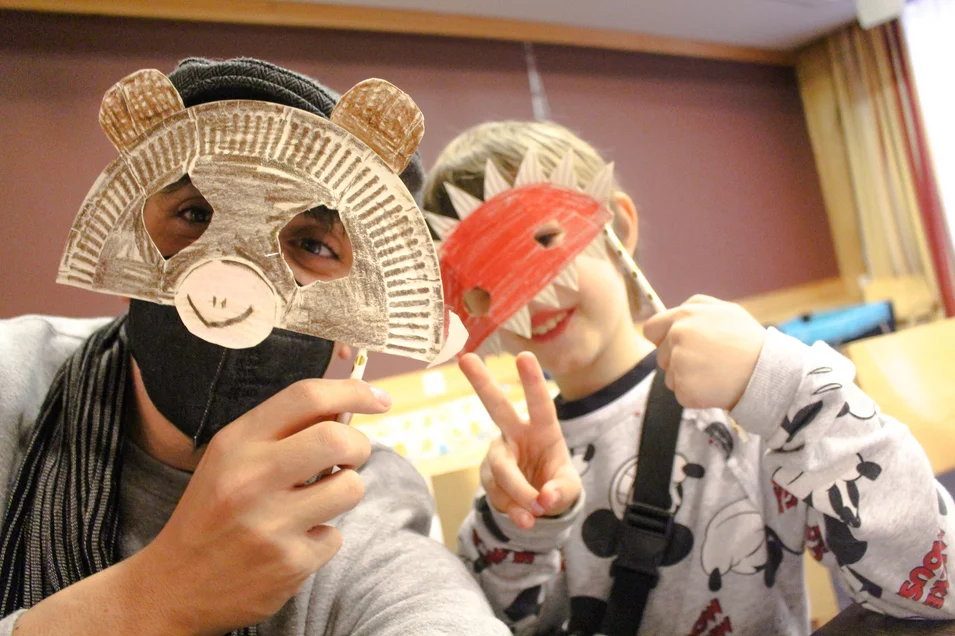 Ehrenamtlicher Mitarbeiter und kleines Mädchen verstecken ihr Gesicht hinter selbst gebastelten Masken
