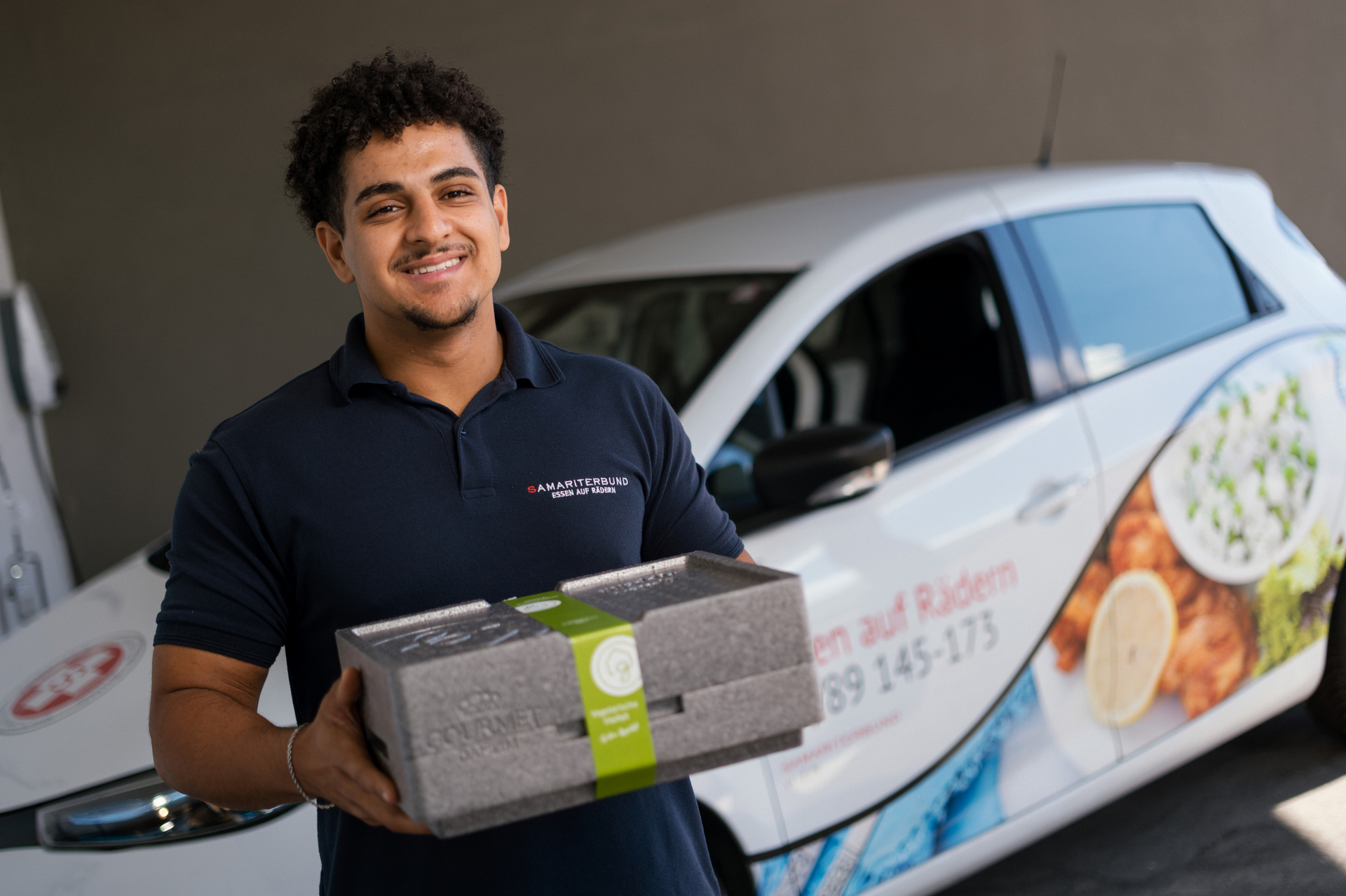 Zivildiener Essen auf Rädern mit Zustellbox vor Essen auf Rädern Auto, lächelt in Kamera