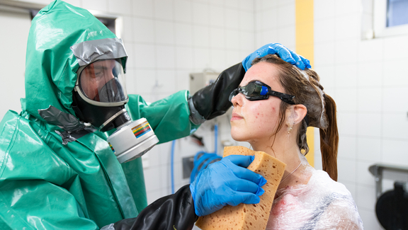 Mann in Schutzanzug mit Atemmaske auf linker Seite, trägt Handschuhe, rechts Frau mit zusammengebundenen Haaren und Schutzbrille, wird von Mann mit einem großen Schwamm abgewischt, im Hintergrund Fliesenwände