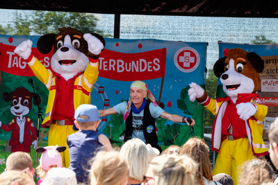 Aufnahme auf Bühne von Tag des Samariterbundes mit Bernhard Fibich und den Maskottchen Sam & Rita auf der Bühne, tanzen und blicken ins Publikum, davor Publikum von hinten, nur Köpfe ersichtlich