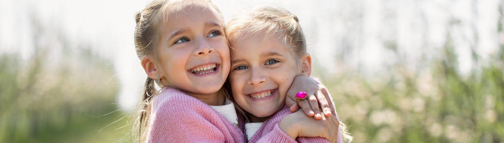 Zwei lächelnde Mädchen umarmen sich in einer blühenden Landschaft. Beide tragen rosa Pullover und haben blonde Haare. Der Hintergrund zeigt eine Reihe von Bäumen, die mit Blüten bedeckt sind. Die Stimmung ist freundlich und fröhlich.