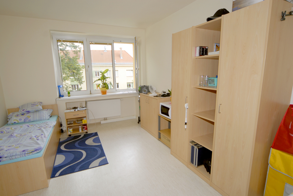 Aufnahme eines Zimmers der Wohnungsloseneinrichtung Haus Sama, links Einzelbett mit Bettwäsche, davor ein Teppich, Fenster mit Blick ins Grüne, Kastenwand mit persönlichen Gegenständen