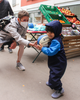 Bezirksvorsteherin 15. Bezirk in Sozialmarkt Pillergasse, kniet Richtung Boden und gibt Kind in Jacke mit Kapuze über dem Kopf Ball, im Hintergrund Kisten mit Obst