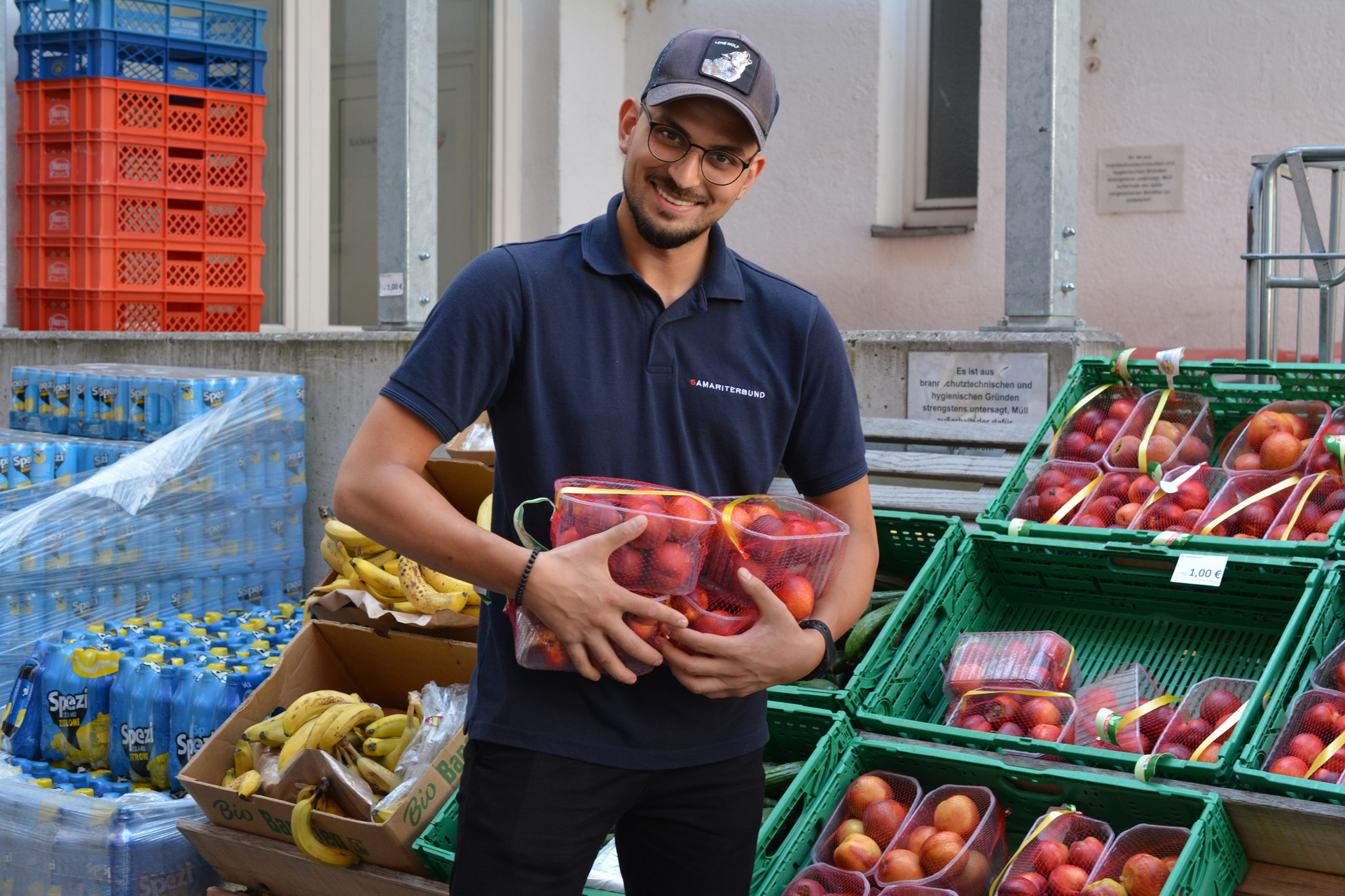 Mitarbeiter im Sozialmarkt mit blauem Samariterbund Polo und Kappe, lächelt in Kamera und hält mehrere Packungen Pfirsiche in den Händen, im Hintergrund weiteres Obst und Gemüse