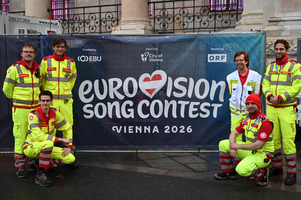 Sechs Rettungskräfte in leuchtenden Uniformen posieren vor einem Banner, das für den Eurovision Song Contest 2026 in Wien wirbt. Das Banner zeigt das Event-Logo und die Gastgeber-Informationen. Die Gruppe steht lächelnd auf einer Straße vor einem historischen Gebäude.