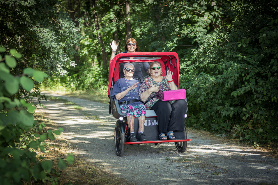 Drei Frauen genießen eine Fahrt in einem Fahrrad-Rikschawagen auf einem Waldweg.