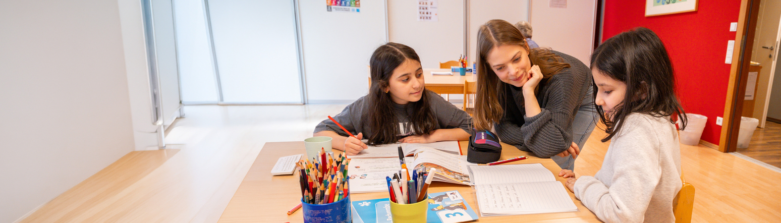 Drei Mädchen arbeiten gemeinsam mit einer Lehrerin an ihren Hausaufgaben am Tisch.