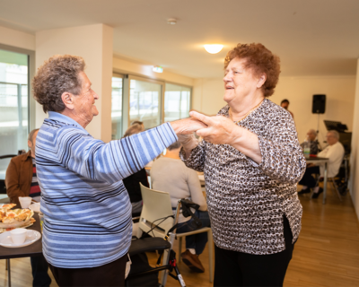 Tag der Senior:innen in der Mühlgrundgasse. Zwei Damen tanzen miteinander, während andere Herrschaften im Hintergrund gemütlich beisammen sitzen.