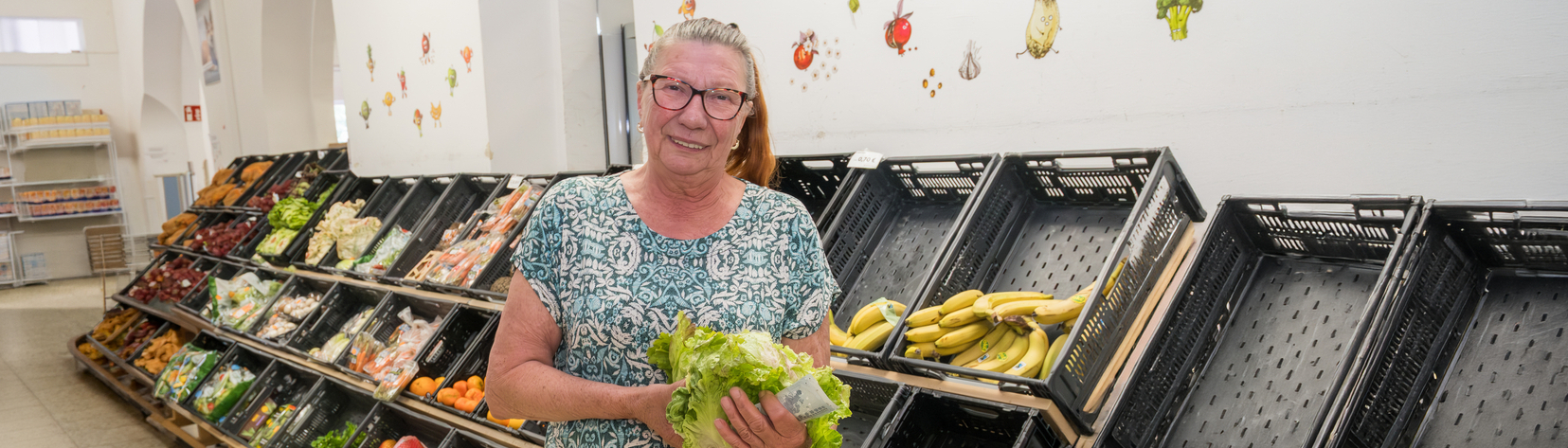 Eine lächelnde Frau steht in einem Raum mit Regalen voller frischer Obst- und Gemüseprodukte. Sie hält einen Bund grüner Blätter in der Hand. Der Hintergrund zeigt gut organisierte Obst- und Gemüsepaletten.