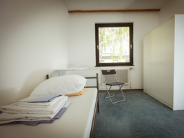 Ein karges Zimmer mit Fenster, Schrank, Stuhl, Tisch und Bett, auf dem Bettwäsche, Kopfkissen, Decke