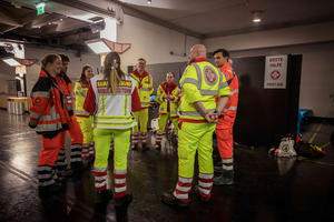 Eine Gruppe von Rettungskräften in auffälliger Schutzkleidung steht in einem Raum. Sie diskutieren im Kreis, während im Hintergrund ein Schild mit der Aufschrift 