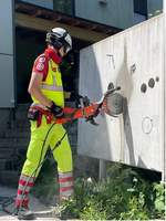 Ein Rettungssanitäter in leuchtend gelber und roter Arbeitsbekleidung schneidet mit einer speziellen Maschine in eine Betonwand. Er trägt einen Helm und schützt sein Gehör. Um ihn herum wachsen grüne Pflanzen und die Umgebung ist hell erleuchtet.