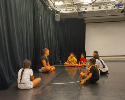 ein Mann sitzt mit ein paar Kindern am Boden in einem Sitzkreis vor einem schwarzen Theatervorhang, LernLEO, Theaterworkshop,  