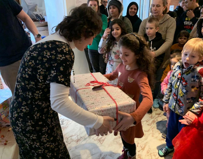 Eine Frau überreicht einem kleinen Mädchen in einem roten Kleid ein weiß verpacktes Geschenk.