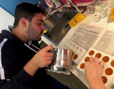 eine Person sitzt an einem Tisch und gießt mit einer Kanne Wachs in Teelichtkerzenhalter, Tisch ist ausgelegt mit Zeitungspapier, im Hintergrund Werkzeug