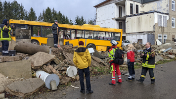 mehrere Personen vor aufgebrochener Straßen, Schäden durch Erdbeben, ein gelber Schulbus auf Schutthaufen, eine Person mit Schaufel auf Schutthaufen, mehrere Personen die dort hin blicken, zwei Personen mit Samariterbund Uniformen