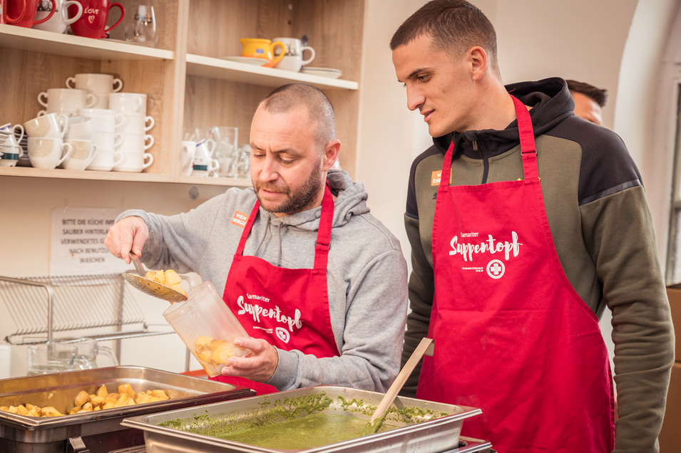 Steffen Hofmann und Nenad Cvetkovic Vertreten den SK Rapid Wien beim Giving Tuesday. Steffen Hofmann füllt gerade einen Plastikbehälter mit Essen, während Nenad Cvetkovic ihm dabei zuschaut.