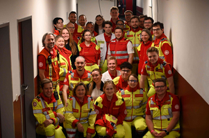Eine Gruppe von etwa 30 Personen steht in einem Flur, viele tragen auffällige gelb-rote Uniformen. Die Mehrheit scheint im Rettungsdienst tätig zu sein. Sie lächeln und posieren gemeinsam für das Foto. Der Hintergrund ist hell und schlicht.
