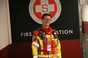 Ein junger Mann in einer auffälligen roten und gelben Warnjacke mit Taschen und Funkgeräten steht vor einem Schild mit dem Schriftzug „FIRST AID STATION“ und einem roten Kreuz. Er lächelt in die Kamera.