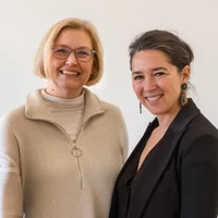 Zwei lächelnde Frauen posieren vor einem hellen Hintergrund.