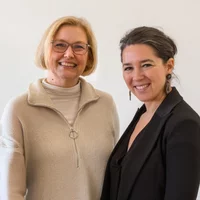 Zwei lächelnde Frauen posieren vor einem hellen Hintergrund.