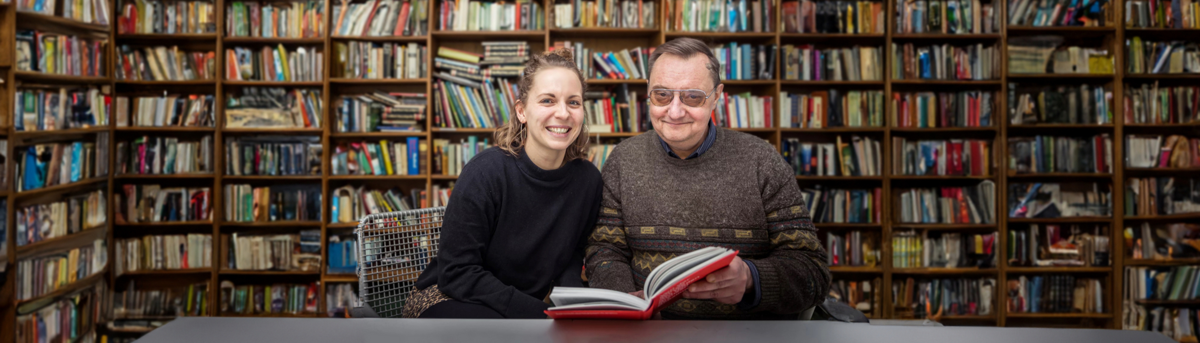 Ein älterer Herr sitz gemeinsam mit einer jüngeren Dame in einer Bibliothek. Sie freuen sich gemeinsam ein Buch zu lesen