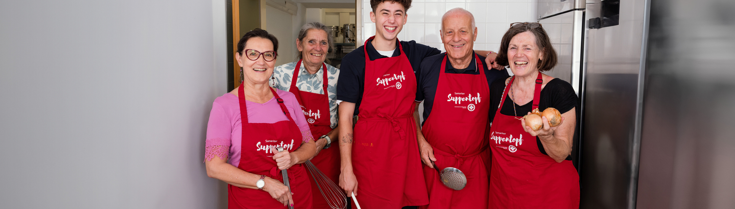 Fünf Personen stehen lächelnd in einer Küche. Sie tragen alle Schürzen mit der Aufschrift 