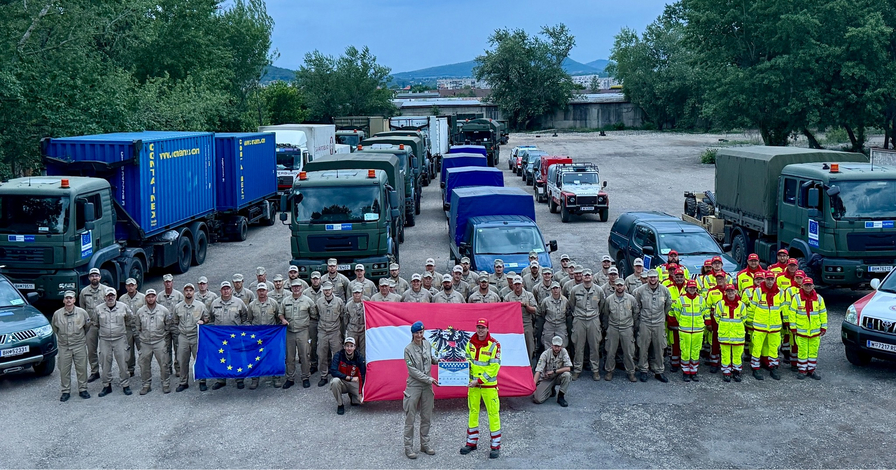 Soldaten und Einsatzkräfte stehen mit Flaggen vor bereitstehenden Fahrzeugen.