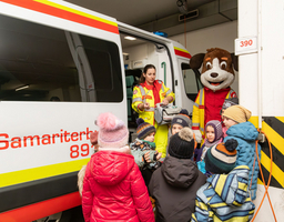 Auf dem Bild ist ein Rettungsauto, dessen Tür geöffnet ist. Vor dem Auto steht eine Gruppe von Kindern, die einer Sanitäterin zuhören. Daneben steht ein Maskottchen.